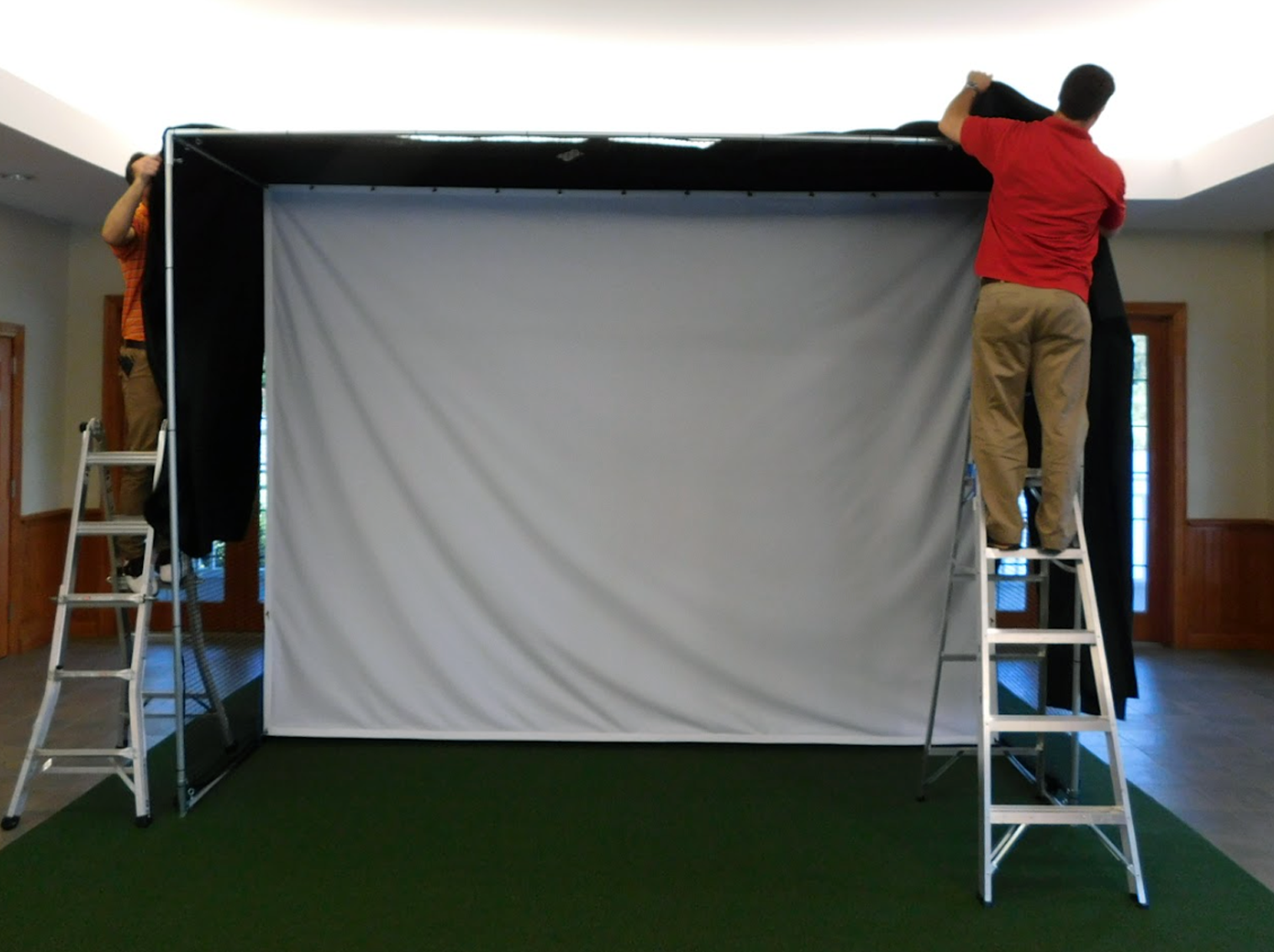 In a room with green carpet, two individuals are skillfully setting up a large white screen encased in a black frame, ideal for an AllSportSystems Golf Simulator Installation Service in the Southeast and Midwest USA.