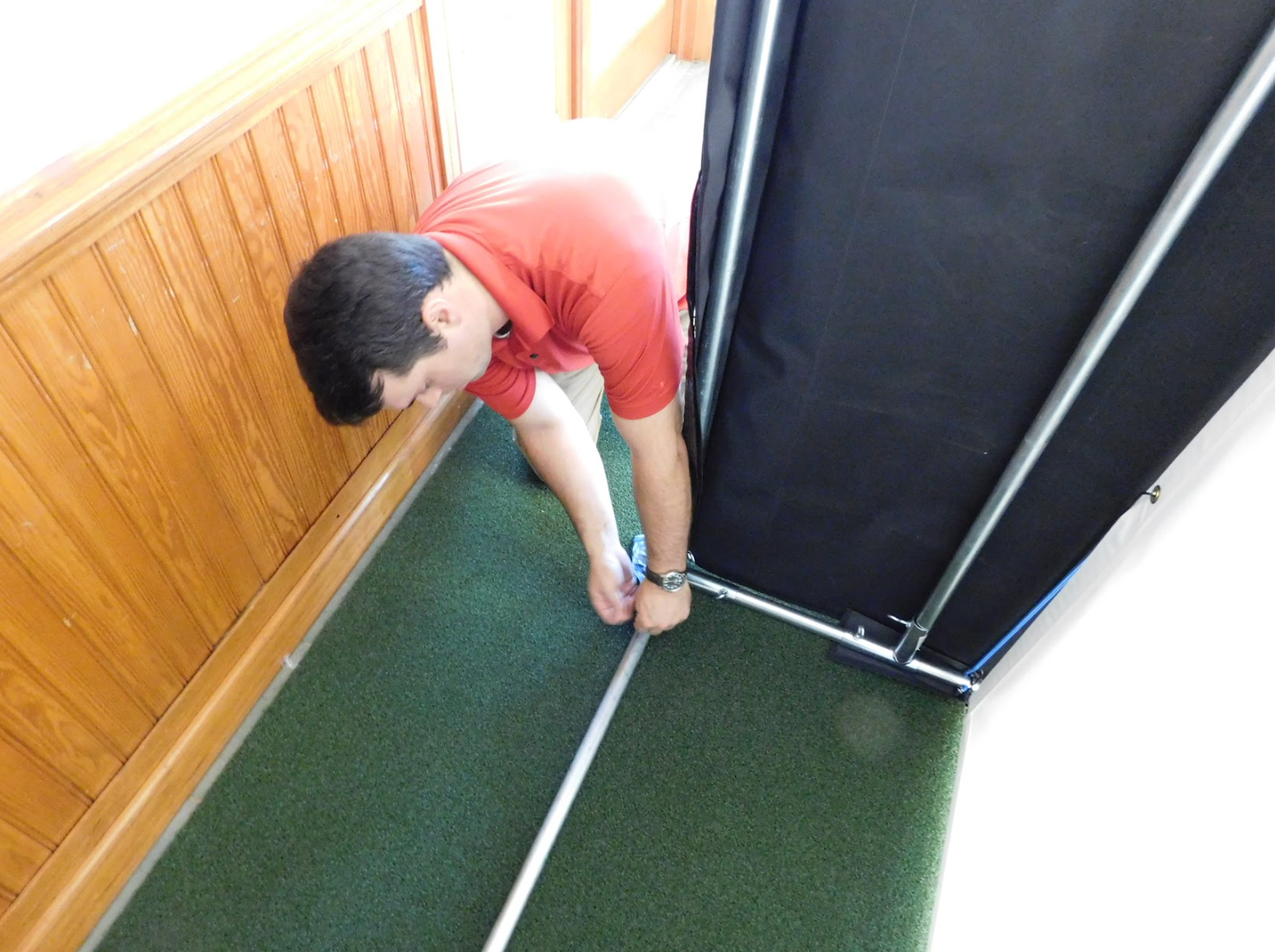A person wearing a red shirt installs a metal frame for an AllSportSystems golf simulator, utilizing Golf Simulator Installation Services in the Southeast and Midwest USA, on a green carpeted floor next to a wooden-paneled wall.