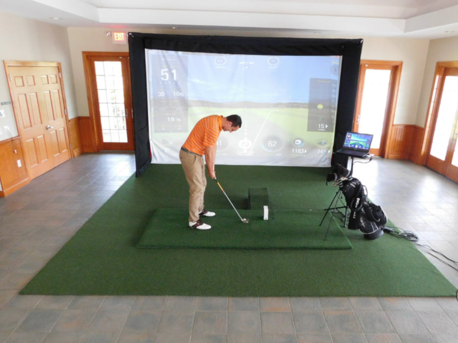 A golfer is practicing their swing on an indoor simulator by AllSportSystems, standing on TurfRoll™ Premium Artificial Turf for Home Golf Simulator Flooring. A virtual golf course is projected on the screen ahead, with golf equipment and a monitor situated close by.