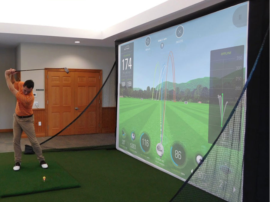 A person is honing their golf swing using the AllSportSystems' indoor simulator equipped with TurfRoll™ Premium Artificial Turf, allowing them to swing a club in front of a large screen that showcases detailed shot data and captivating virtual course visuals.