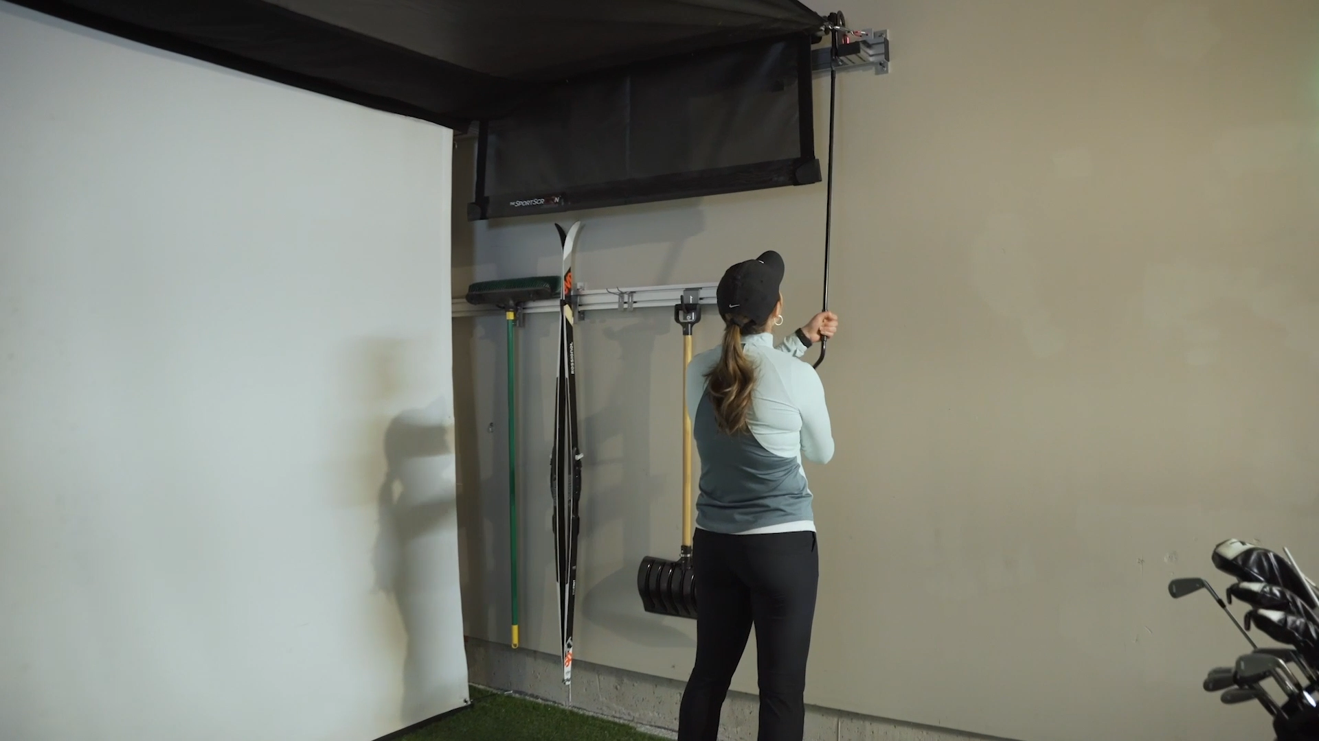 A person adjusts an AllSportSystems SportScreen Vanish Retractable Golf Bay by a wall in their home gym, with golf equipment nearby.