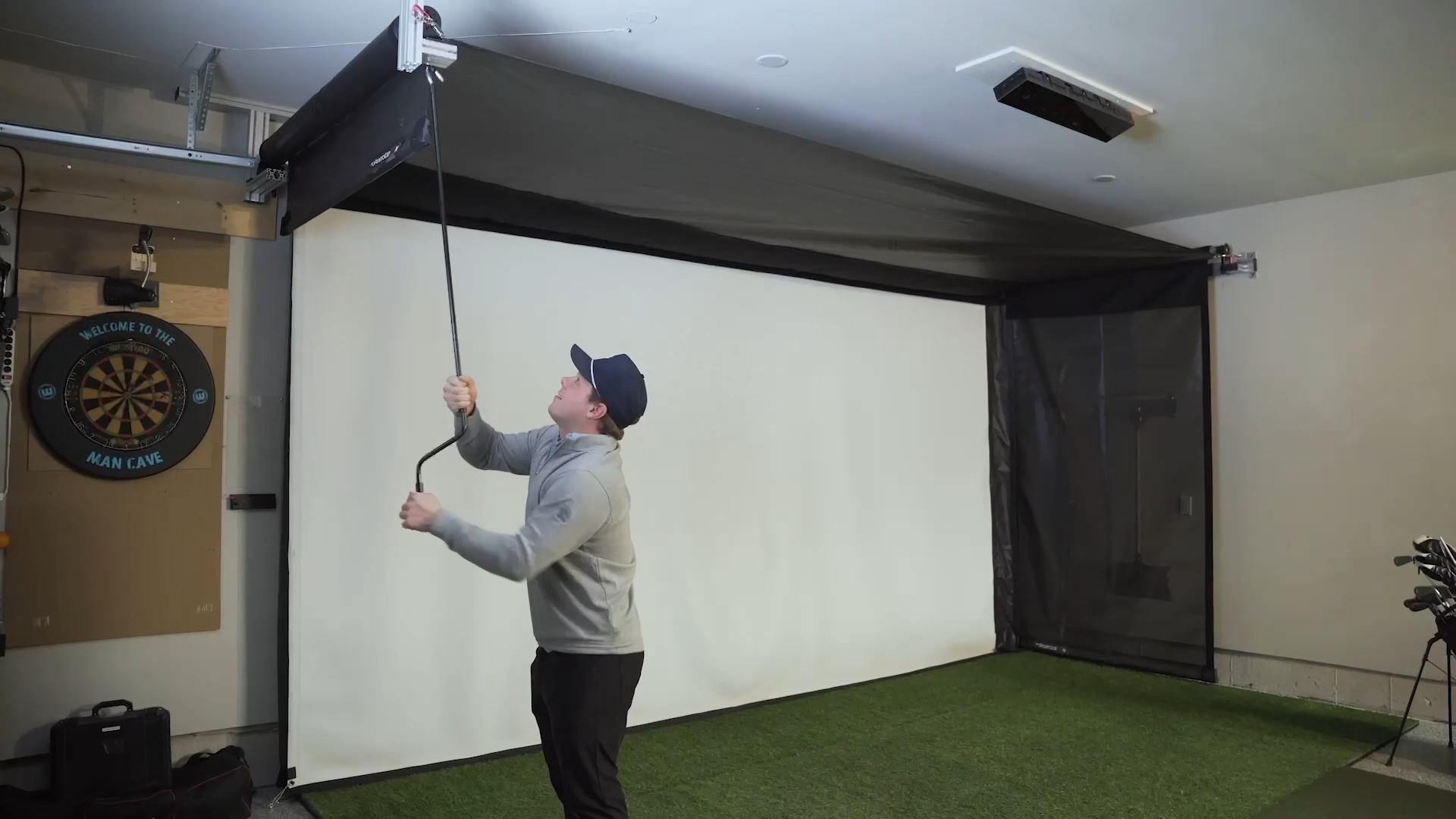 A person in a gray sweater and cap adjusts a ceiling-mounted projector in a room with an AllSportSystems SportScreen Vanish Retractable Golf Bays and Enclosures, alongside a dartboard and golf putting mat.