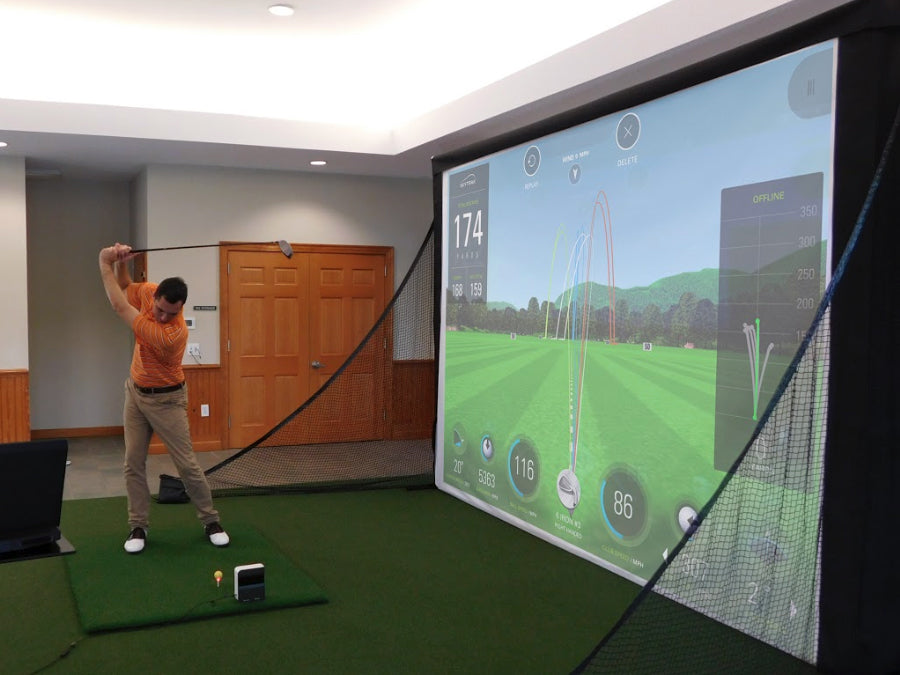 A person practices their golf swing indoors using the Fast Bundle Complete Uneekor Eye XO Kit with a 4K projector and bay, while a screen displays swing data and distance.