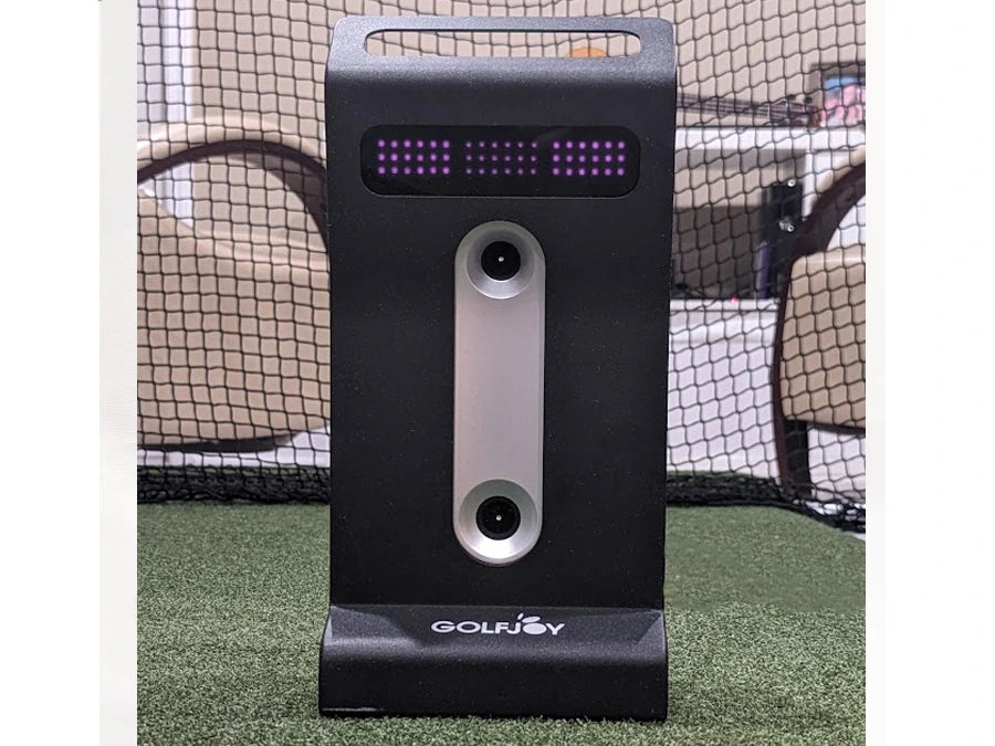 A black GolfJoy GDS Plus Launch Monitor with screen and cameras stands on turf. Compatible with GDS Plus golf simulator, it enhances practice sessions. A net and chairs in the background complete the immersive golfing setup.